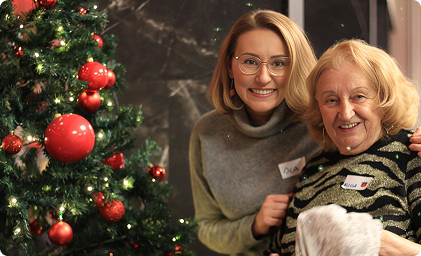 Wolontariuszka i seniorka uśmiechają się do zdjęcia obok choinki.